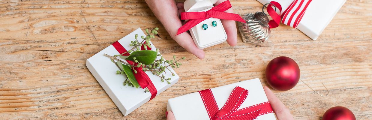 Christmas themed gift wrapped square boxes, on person holds emeral square earrings in white jewelry box with a ribbon 